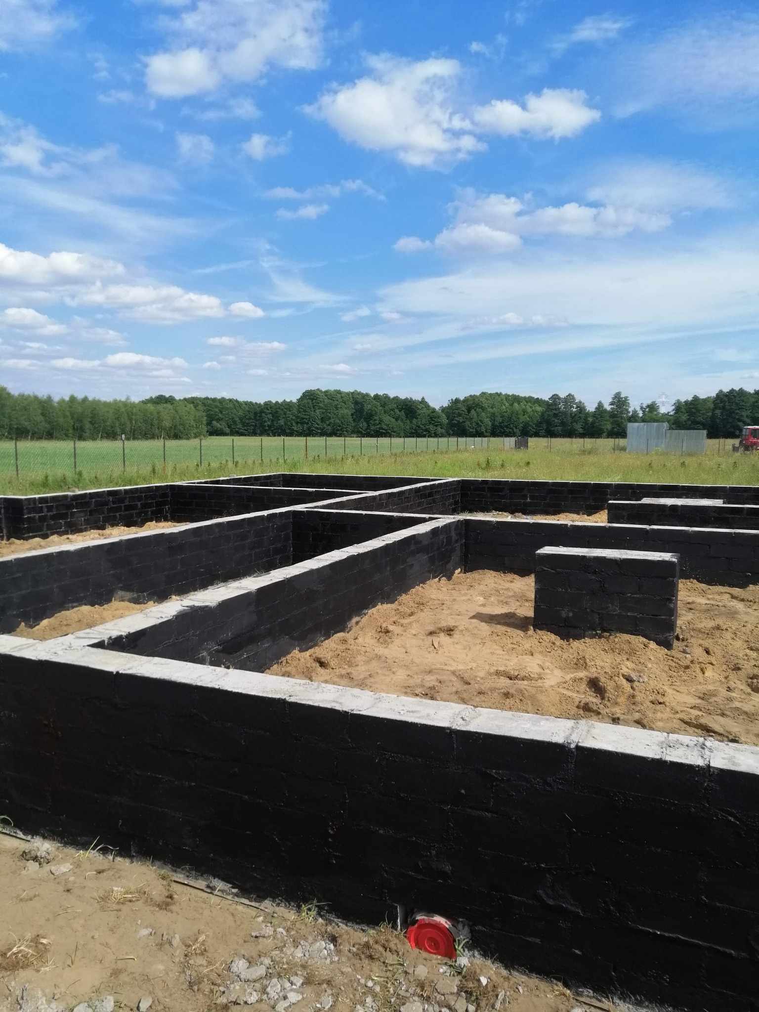 Zabezpieczone hydroizolacją fundamenty z bloczków betonowych, wypełnione piaskiem, na tle zieleni i błękitnego nieba z chmurami.