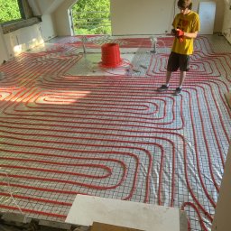 Install Home KONRAD ZOWCZAK - Czerwone rury ogrzewania podłogowego ułożone na siatce w jasnym pomieszczeniu poddasza, obok stoi instalator w żółtej koszulce i czerwonych rękawicach.