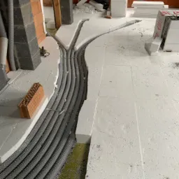 Szare rury w bruzdach izolacji podłogowej, rozchodzące się w dwóch kierunkach. W tle cegły i styropian. Widok z góry.