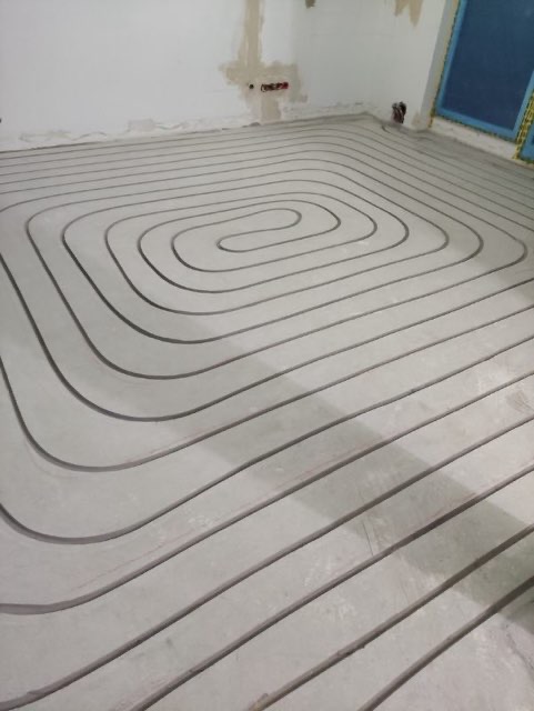 Spiralny układ rur ogrzewania podłogowego zalanych betonem w surowym wnętrzu. Widoczne ślady po instalacjach na ścianie i fragment okna z niebieską ramą.