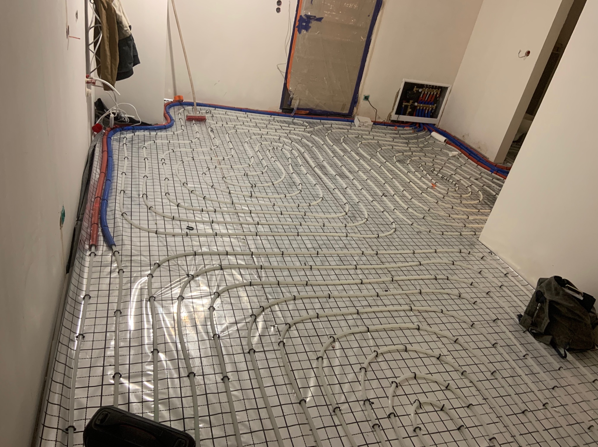 Białe rury ogrzewania podłogowego ułożone na folii z siatką w surowym wnętrzu. Na ścianie widoczna rozdzielnica. Częściowo widoczny worek z narzędziami.
