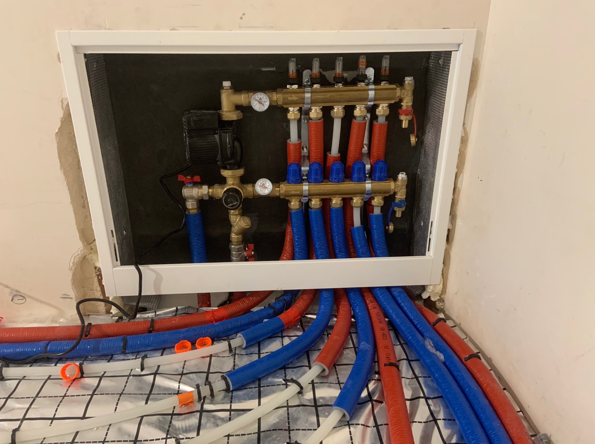 Rozdzielacz ogrzewania podłogowego z rurami w kolorze niebieskim i czerwonym, zamontowany na folii aluminiowej z siatką, umieszczony w białej obudowie.