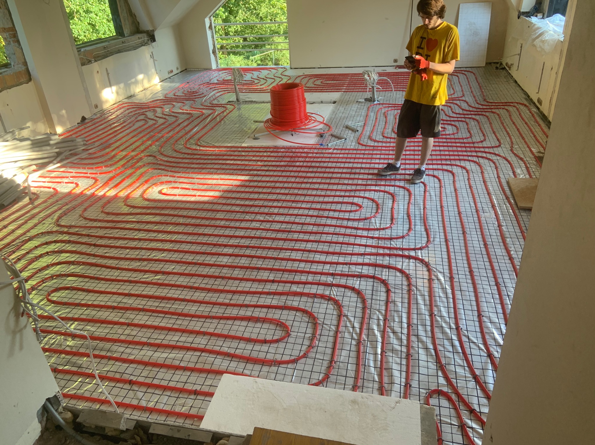 Czerwone rury ogrzewania podłogowego ułożone na siatce w jasnym pomieszczeniu poddasza, obok stoi instalator w żółtej koszulce i czerwonych rękawicach.
