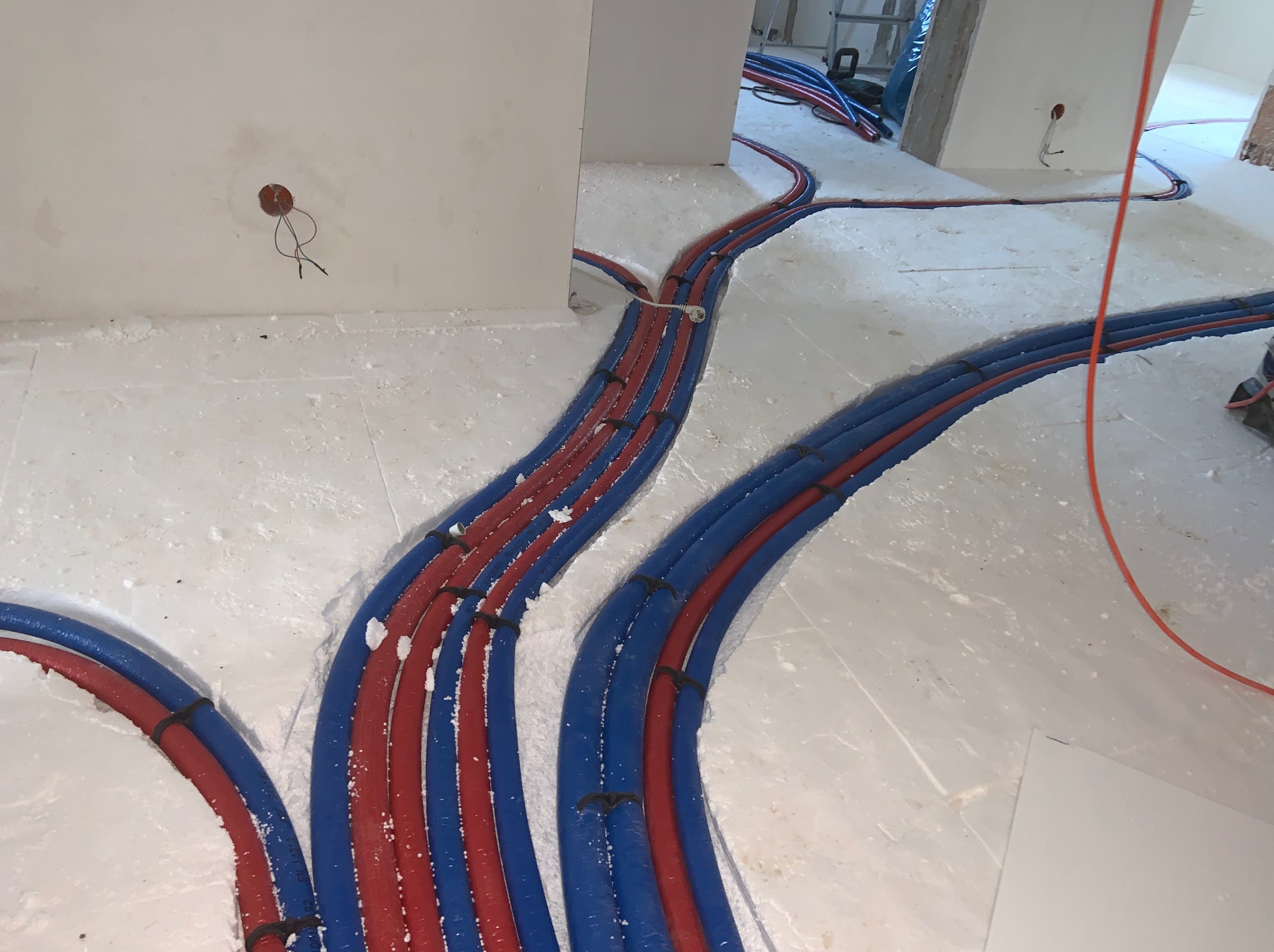 Rozłożone rury ogrzewania podłogowego w kolorach czerwonym i niebieskim, przymocowane do podłoża. Widoczne fragmenty ścian i wystające kable elektryczne.