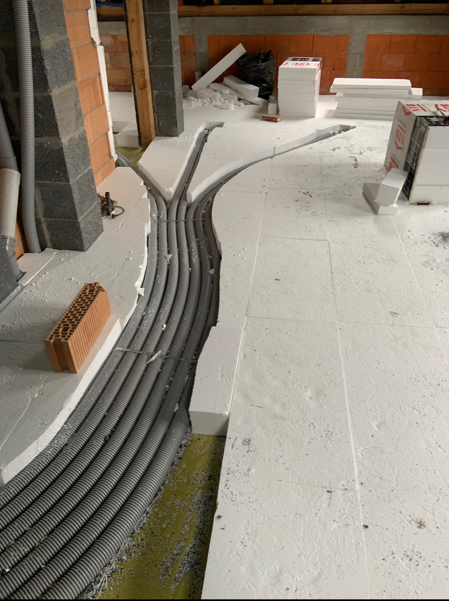 Szare rury w bruzdach izolacji podłogowej, rozchodzące się w dwóch kierunkach. W tle cegły i styropian. Widok z góry.