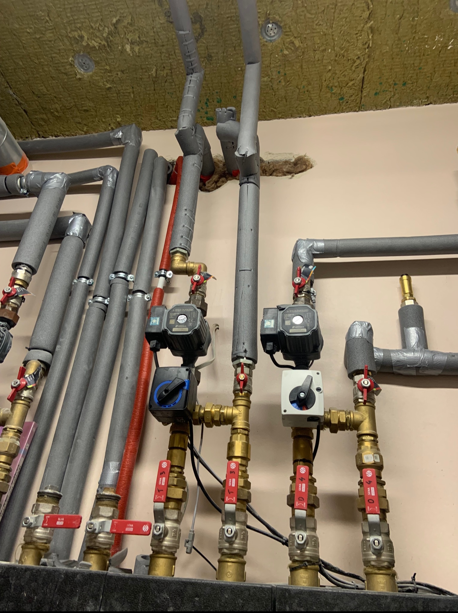 Szczegółowe ujęcie instalacji centralnego ogrzewania z izolowanymi rurami, zaworami z czerwonymi uchwytami i pompami obiegowymi, zamontowanej pod sufitem z wełny mineralnej.