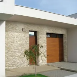 Nowoczesny dom z elewacją z kamienia dekoracyjnego Incana, brązowe drzwi i brama garażowa, oraz młode drzewko na trawniku.