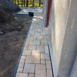 Ułożona ścieżka z kostki brukowej w odcieniach beżu i szarości, przylegająca do ściany budynku, z widocznym przygotowanym podłożem i stertą ziemi w tle.