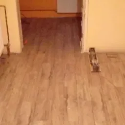 Ułożona drewniana podłoga w pokoju z żółtymi ścianami, w tle widoczne okno i grzejnik, przy ścianie leży narzędzie do cięcia listew przypodłogowych.