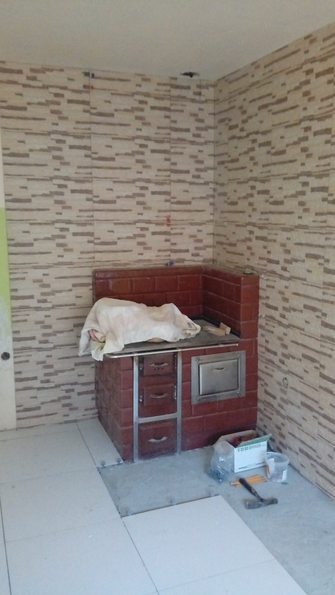 Wykończona ściana z beżowo-brązowych płytek w trakcie prac wykończeniowych, piec kaflowy częściowo zakryty materiałem ochronnym, na podłodze białe płytki, brakuje kilku elementów, widoczne...