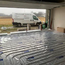 Wnętrze garażu z ułożoną instalacją ogrzewania podłogowego na folii z nadrukiem, widoczny biały bus firmy instalacyjnej na zewnątrz, szara pogoda.