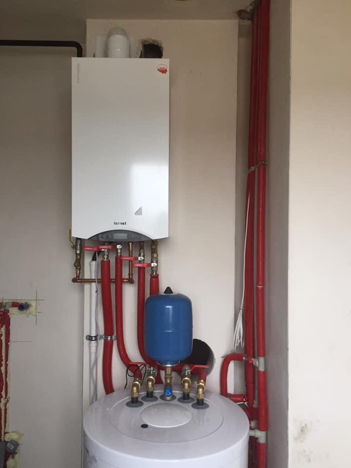 Biały, wiszący piec gazowy marki Termet, zainstalowany nad białym zasobnikiem wody z niebieskim naczyniem przeponowym, widoczne czerwone rury instalacji grzewczej i zawory, ściany w odcieniach beżu.