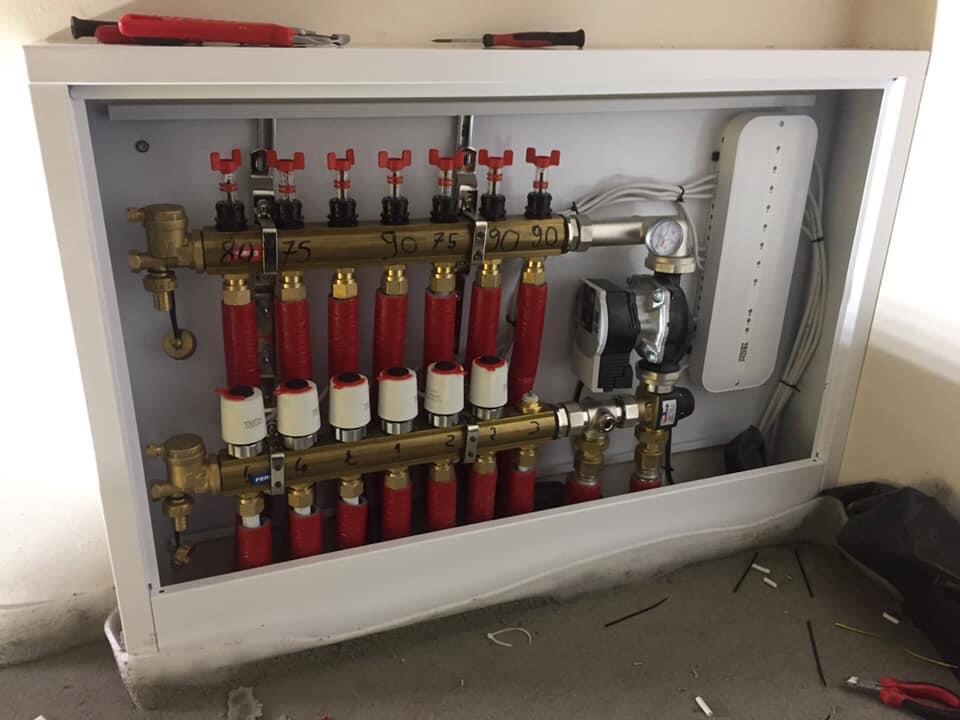 Rozdzielacz ogrzewania podłogowego w białej szafce z widocznymi rurami w izolacji, zaworami termostatycznymi i pompą obiegową, zamontowany na tle surowego betonu.