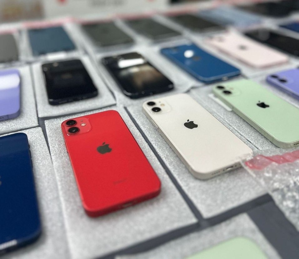 Różnokolorowe telefony iPhone 12 ułożone na szarych, miękkich podkładkach, widoczne w różnych kolorach: czerwonym, białym, zielonym, niebieskim i czarnym.