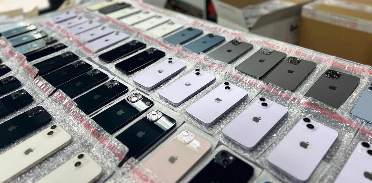 Rząd nowych telefonów iPhone w różnych kolorach, zabezpieczonych folią bąbelkową, ułożonych na stole przed wysyłką lub naprawą.