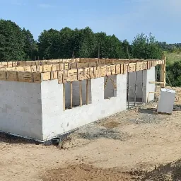 Wznoszenie ścian parterowego domu z bloczków betonowych, widoczne szalunki na strop, w tle zielony krajobraz.