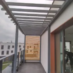 Zadaszenie tarasu z drewnianą pergolą w nowoczesnym stylu, widok z balkonu na budynek mieszkalny, pracownik na drabinie w tle.