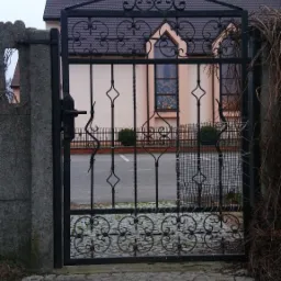 Kuta, czarna brama z ozdobnymi elementami na posesji z widocznym budynkiem w tle, widok z brukowanego podjazdu.