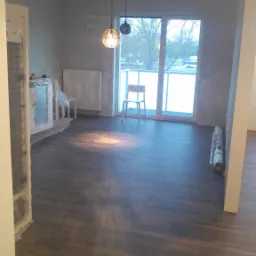 Pokój po remoncie z nową, ciemną podłogą z paneli, widoczne dwie lampy wiszące, okno balkonowe i meble zabezpieczone folią malarską.