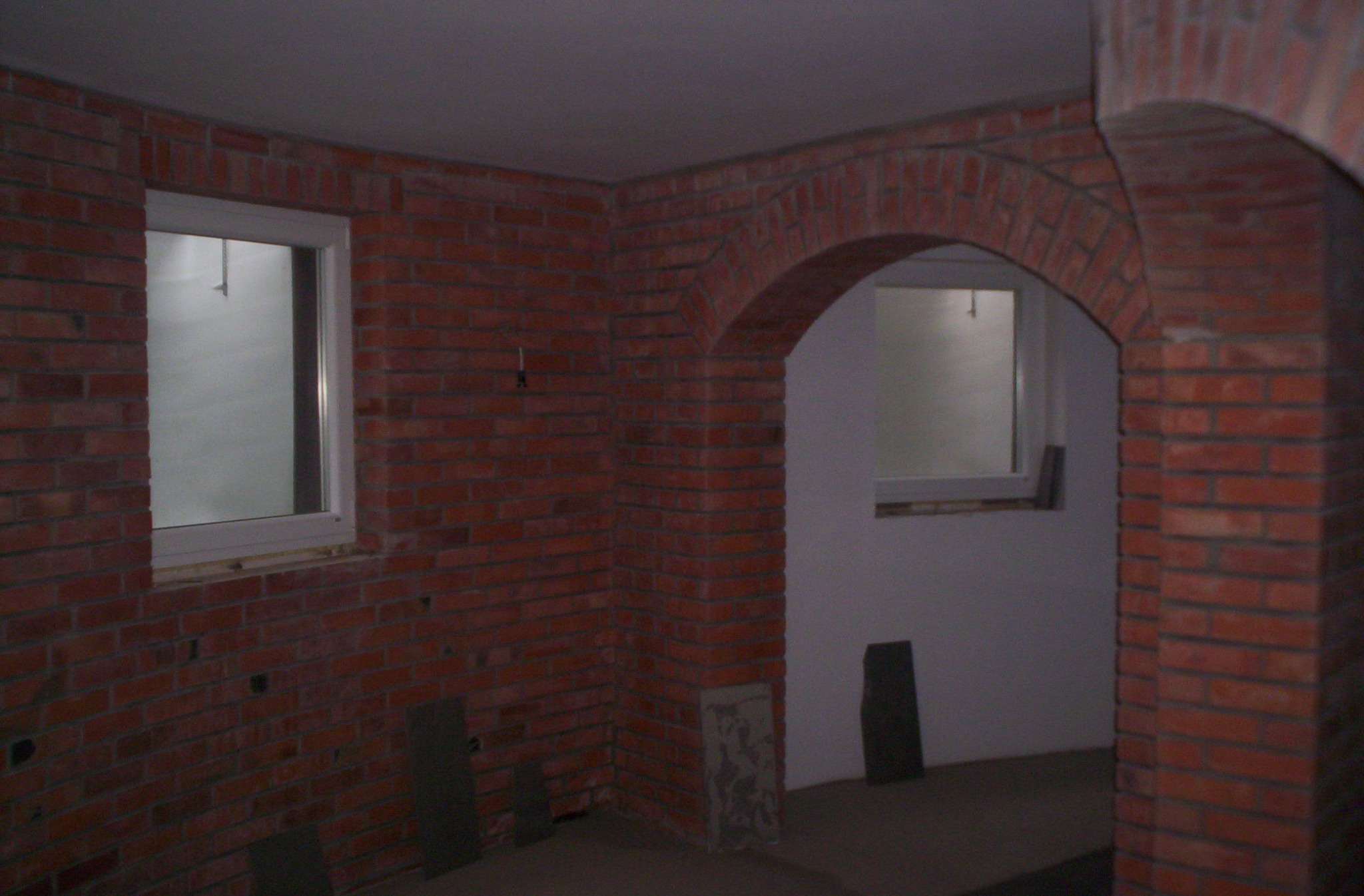 Surowa ściana z czerwonej cegły z oknem i łukowym przejściem, widoczne ślady po instalacjach elektrycznych, na podłodze płyty