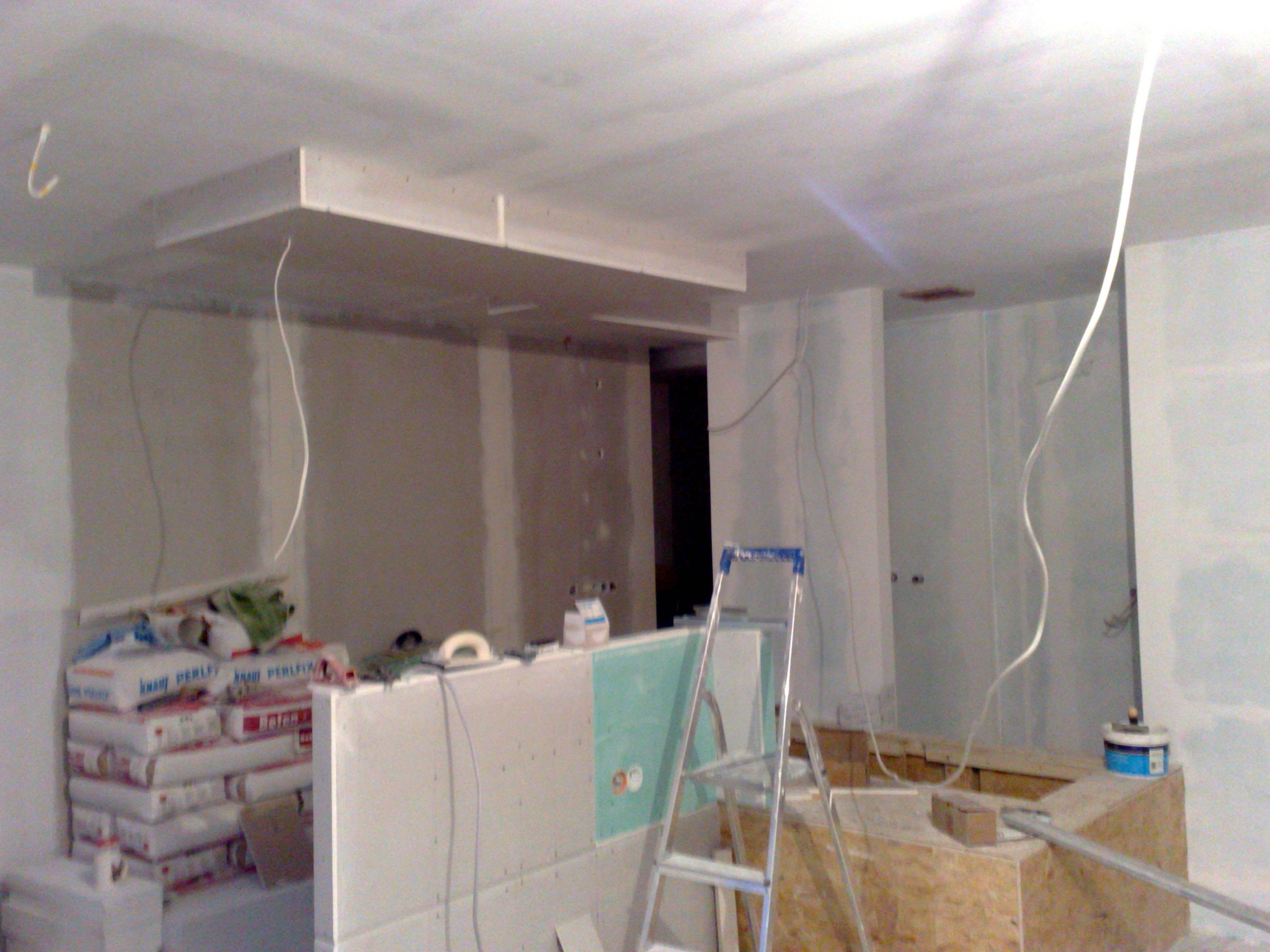 Wnętrze w trakcie prac wykończeniowych: widoczne płyty gipsowo-kartonowe na ścianach i suficie, drabina aluminiowa, worki z zaprawą, instalacja elektryczna w trakcie montażu.