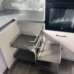 Wysuwane półki cargo w szafce narożnej w nowoczesnej kuchni z szarym blatem i marmurową ścianą nad blatem, obok zabudowany piekarnik marki Beko.