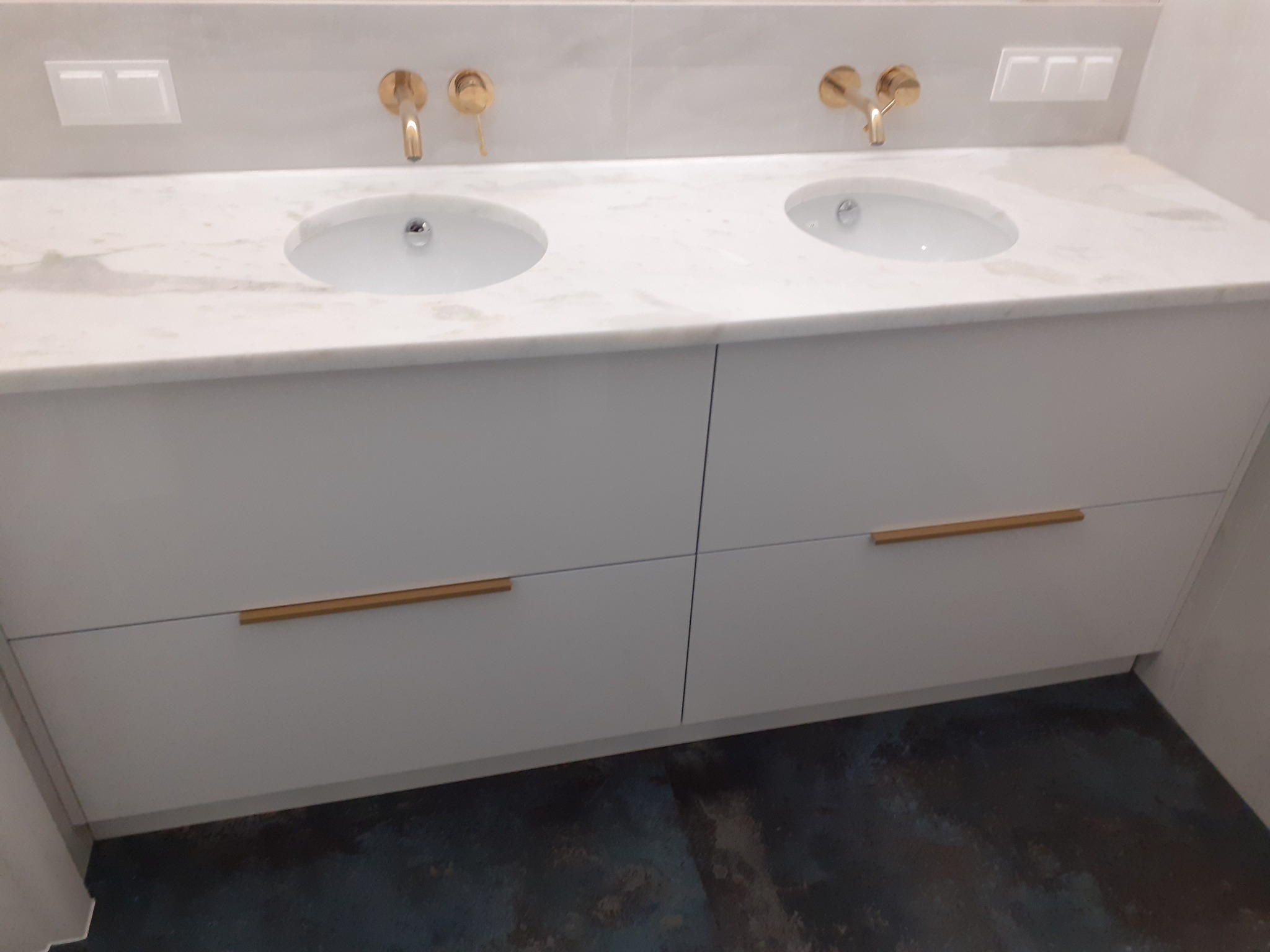 Biała szafka łazienkowa pod umywalki z marmurowym blatem i złotymi bateriami, widoczne dwa okrągłe umywalki i minimalistyczne uchwyty.