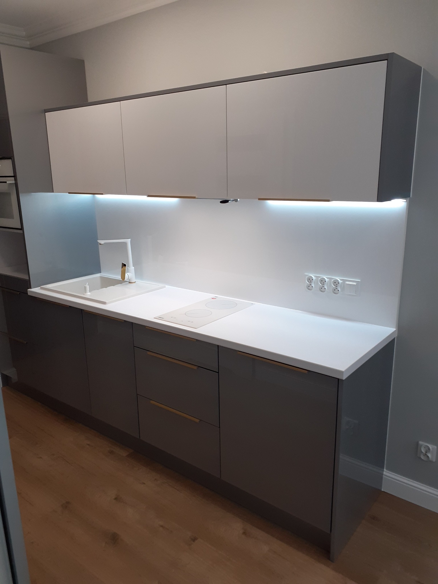 Nowoczesna kuchnia w odcieniach szarości z białym blatem i frontami szafek górnych, podświetleniem LED pod szafkami wiszącymi i złotymi akcentami w postaci uchwytów oraz baterii.