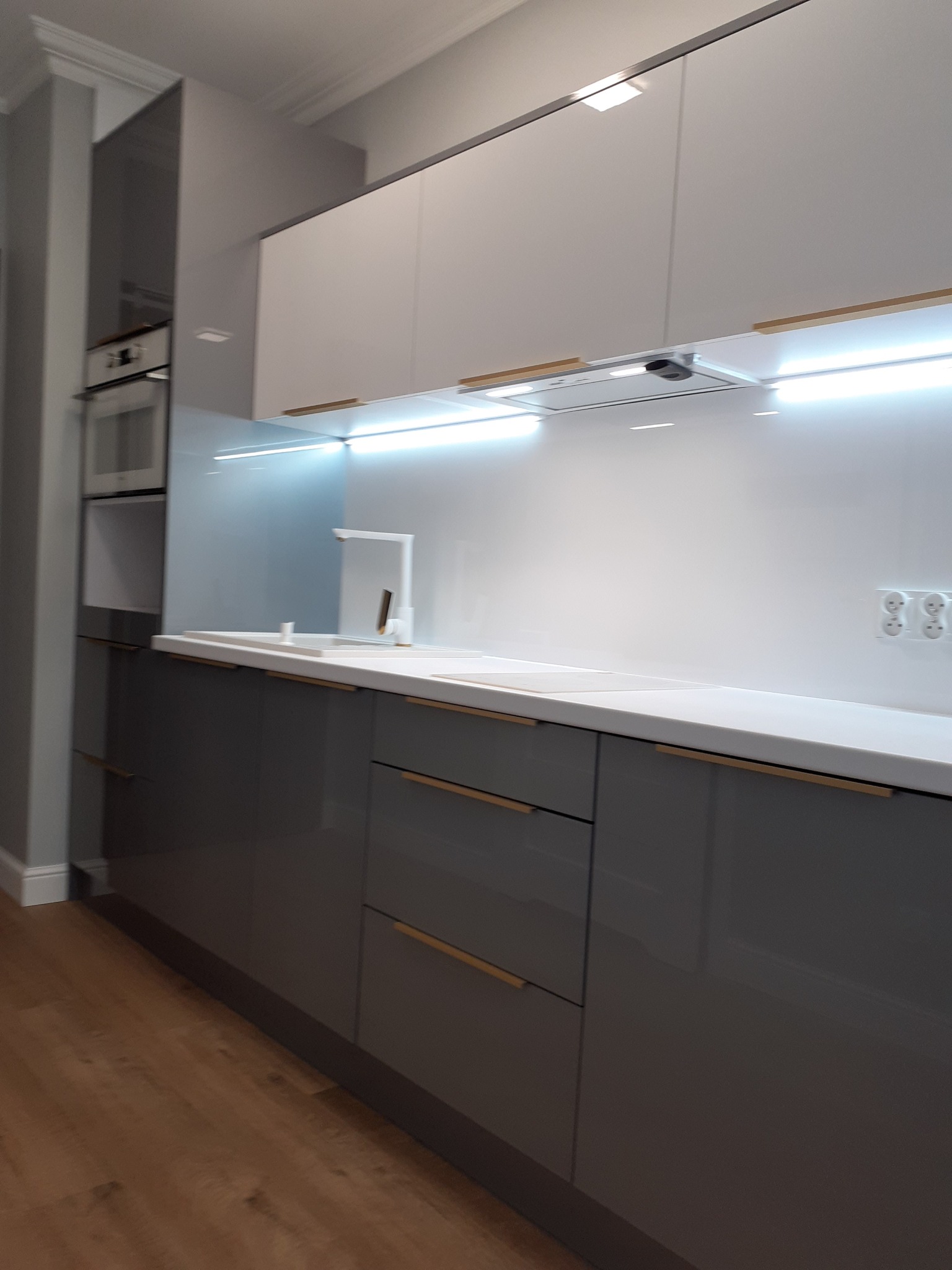 Nowoczesna kuchnia z szarymi, lakierowanymi szafkami dolnymi i białymi górnymi, podświetlona listwą LED pod szafkami wiszącymi, z drewnianymi akcentami na uchwytach.