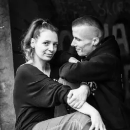 Czarno-białe zdjęcie pary: kobieta z włosami spiętymi w kucyk patrzy w obiektyw, mężczyzna z kr&oacute;tkimi włosami obejmuje ją, oboje siedzą na drewnianej powierzchni na tle graffiti.