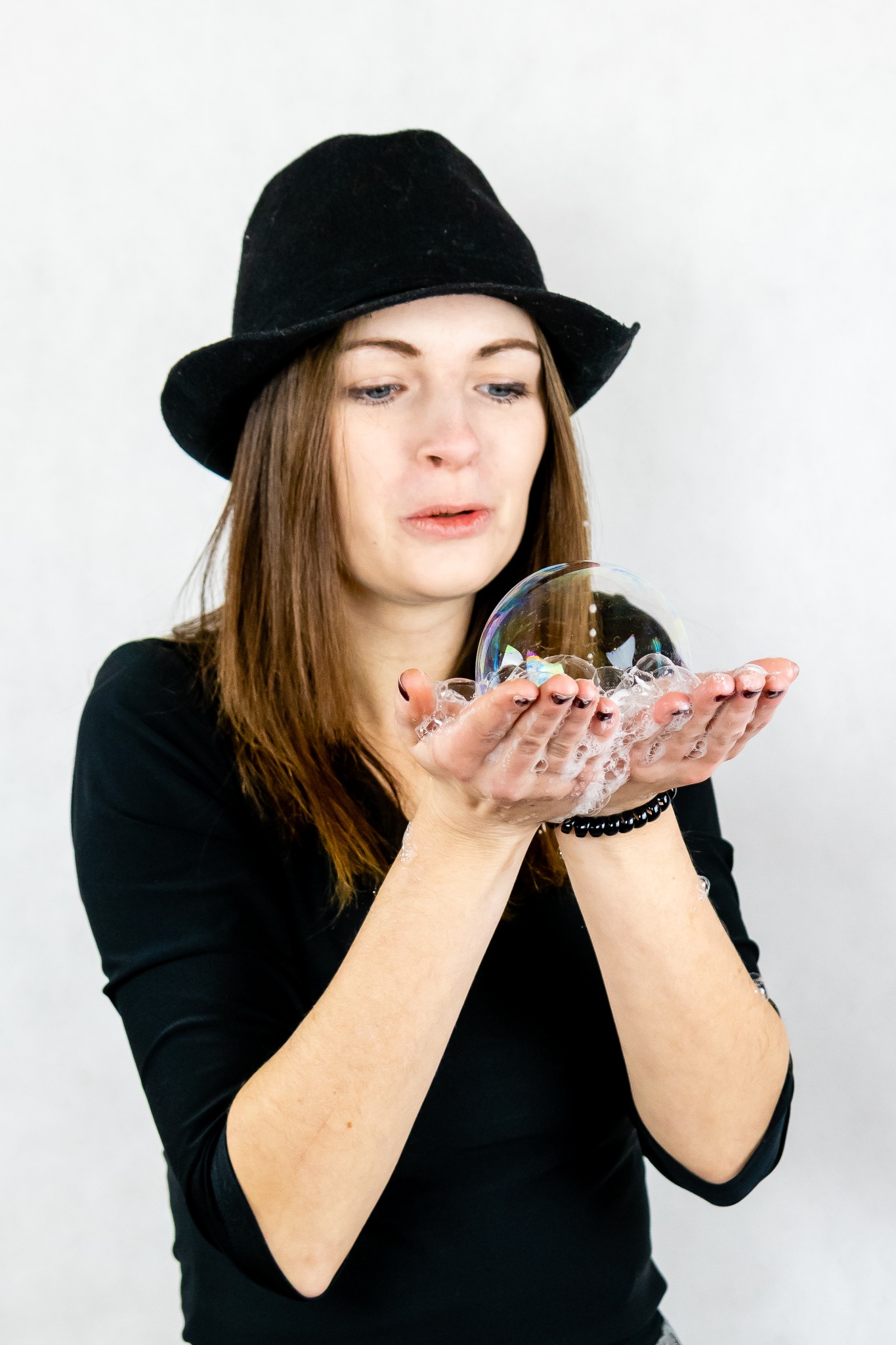 Kobieta w czarnym kapeluszu dmucha bańki mydlane, trzymając je w dłoniach z widoczną bransoletką.