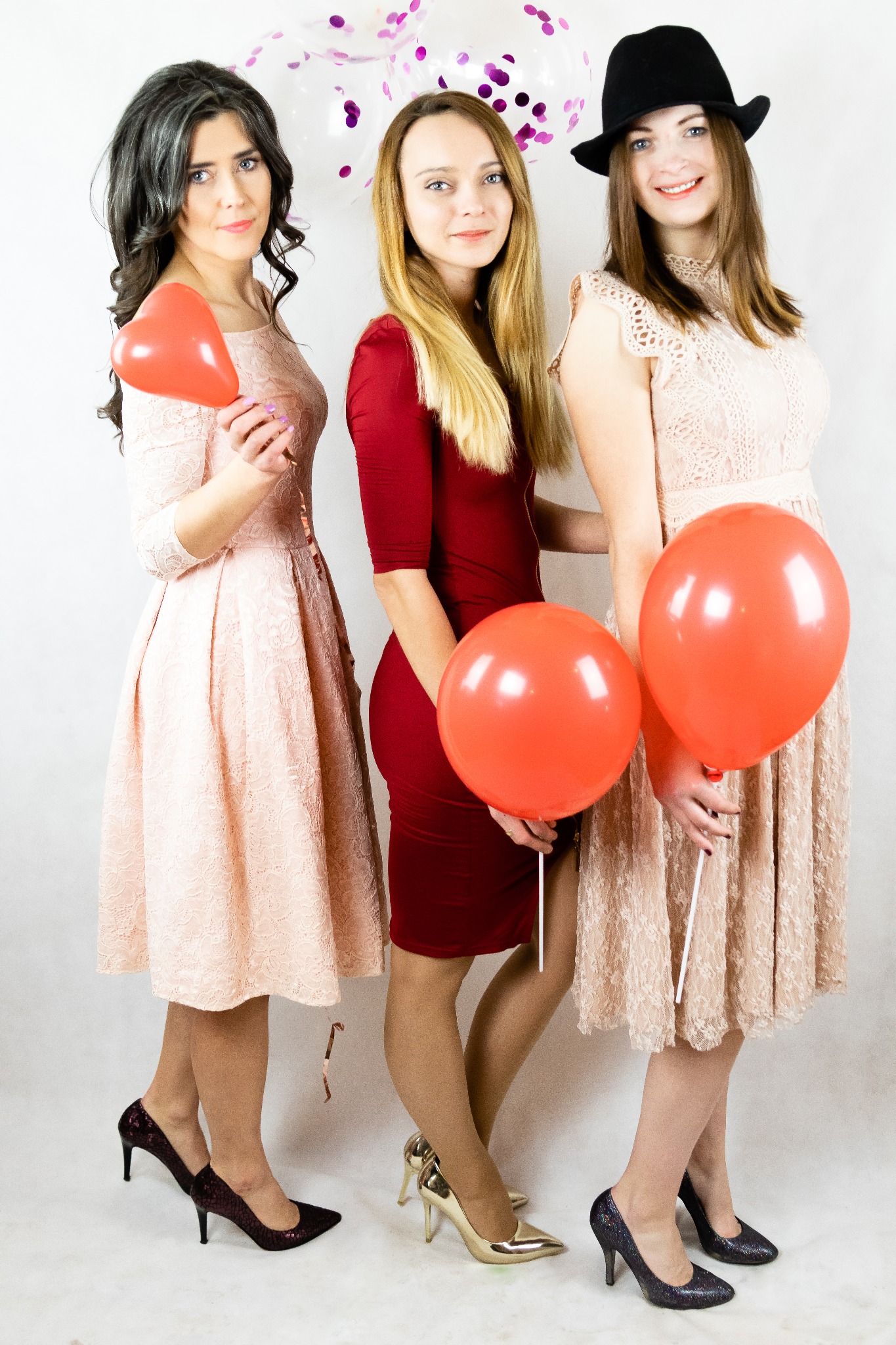 Trzy uśmiechnięte kobiety w eleganckich sukienkach pozują z czerwonymi balonami na tle białych balonów z konfetti.