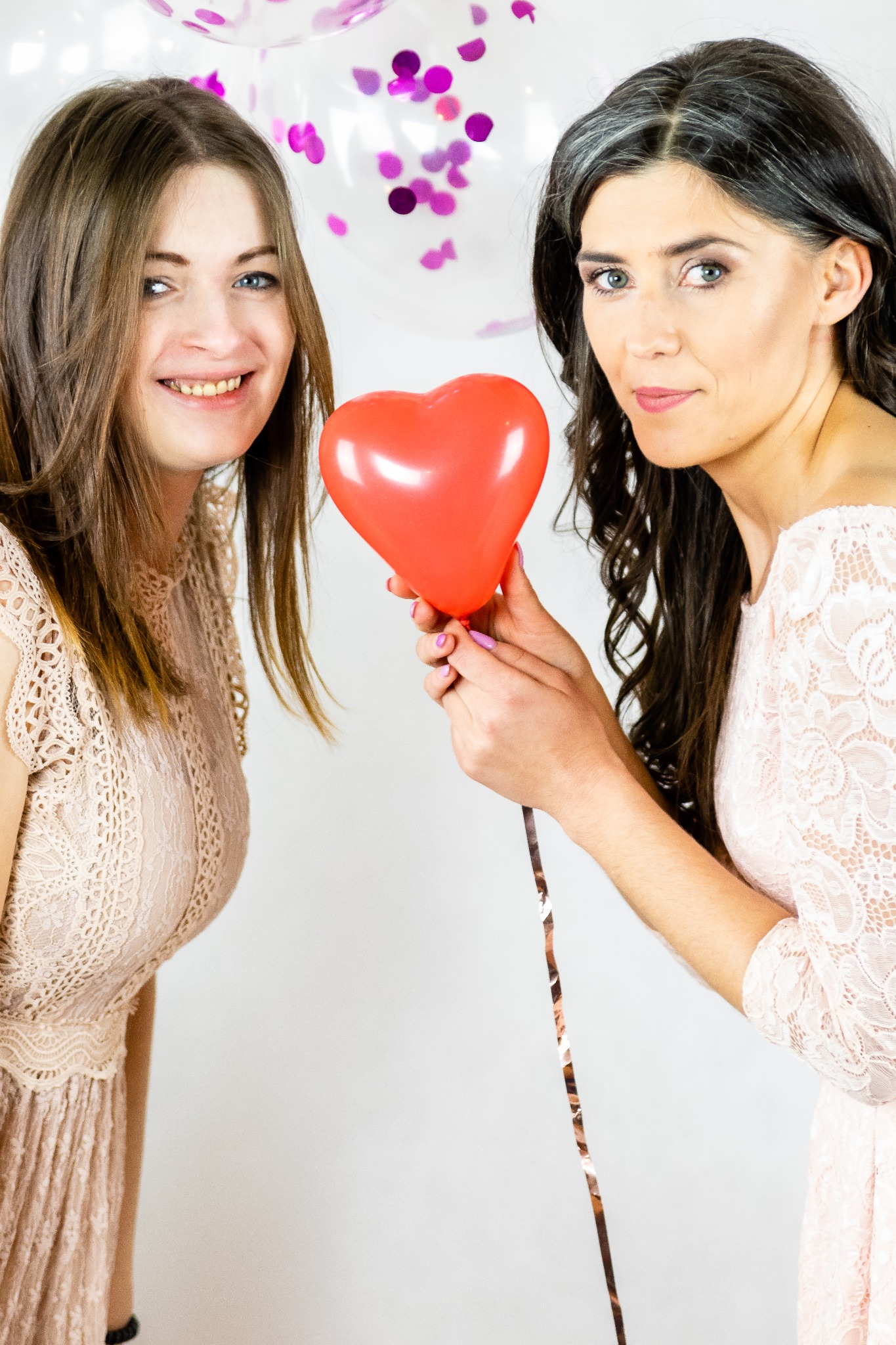 Dwie kobiety w koronkowych sukienkach trzymają razem czerwony balon w kształcie serca na tle białych balonów z konfetti w odcieniach fioletu.