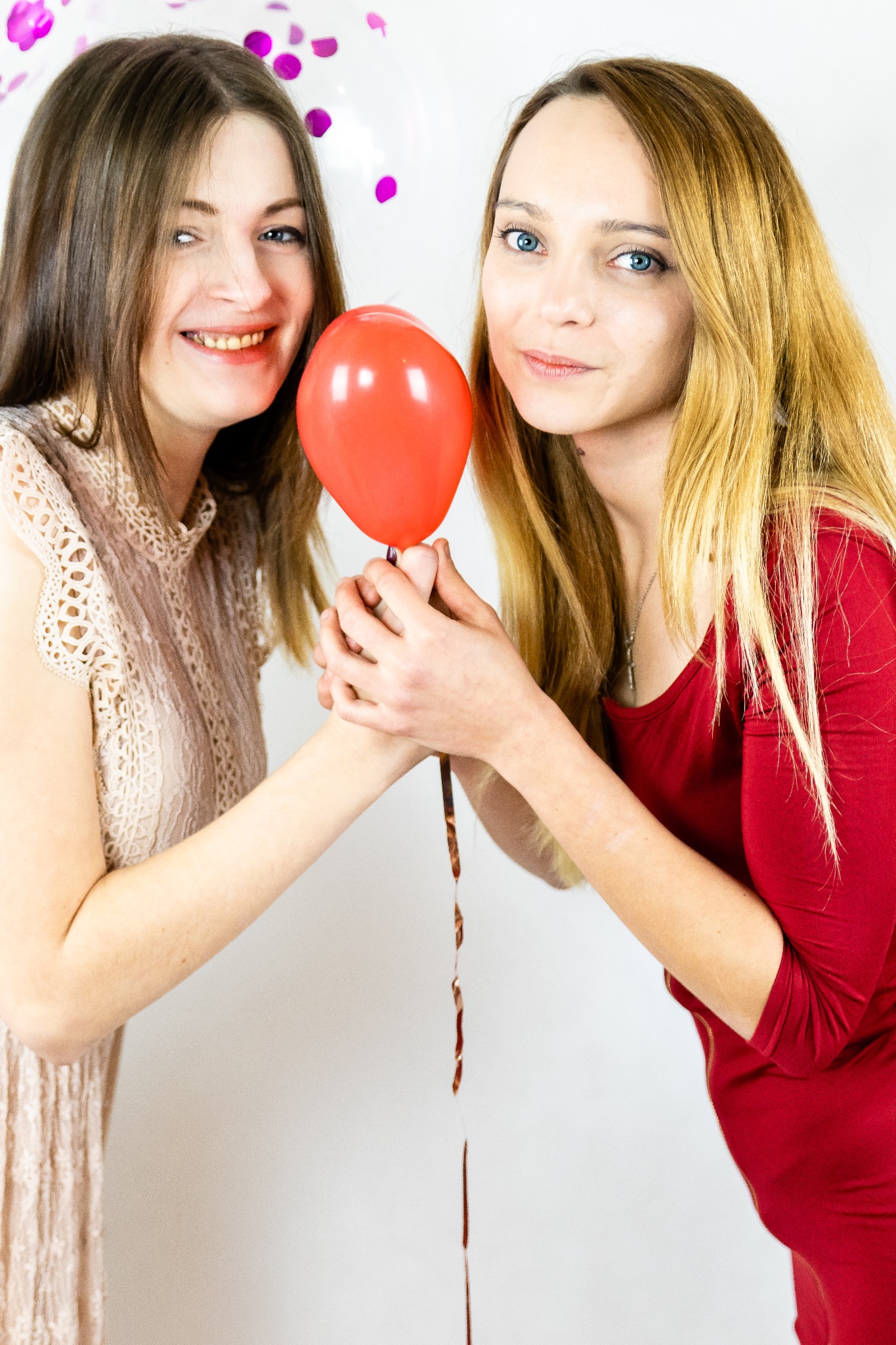 Dwie uśmiechnięte kobiety trzymają razem czerwony balon na tle białej ściany z konfetti.