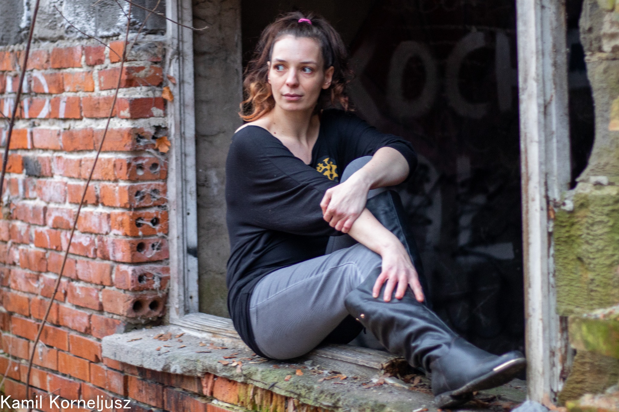 Kobieta w czarnej bluzce i kozakach pozuje siedząc w zniszczonym oknie z ceglaną ramą, na ścianie za nią widoczne graffiti.
