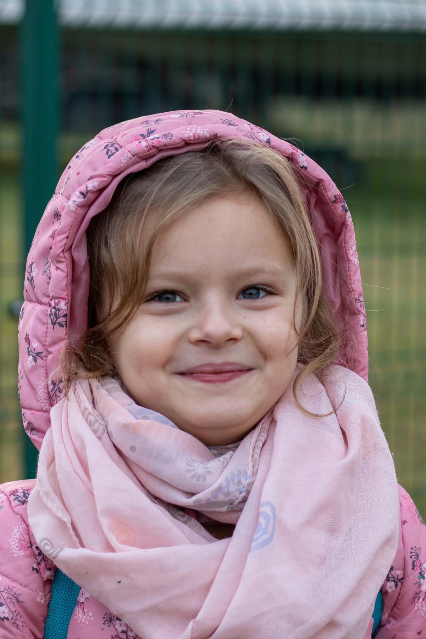 Portret uśmiechniętej dziewczynki z niebieskimi oczami w różowej, kwiecistej kurtce z kapturem i jasnoróżowym szaliku na tle zielonego ogrodzenia.