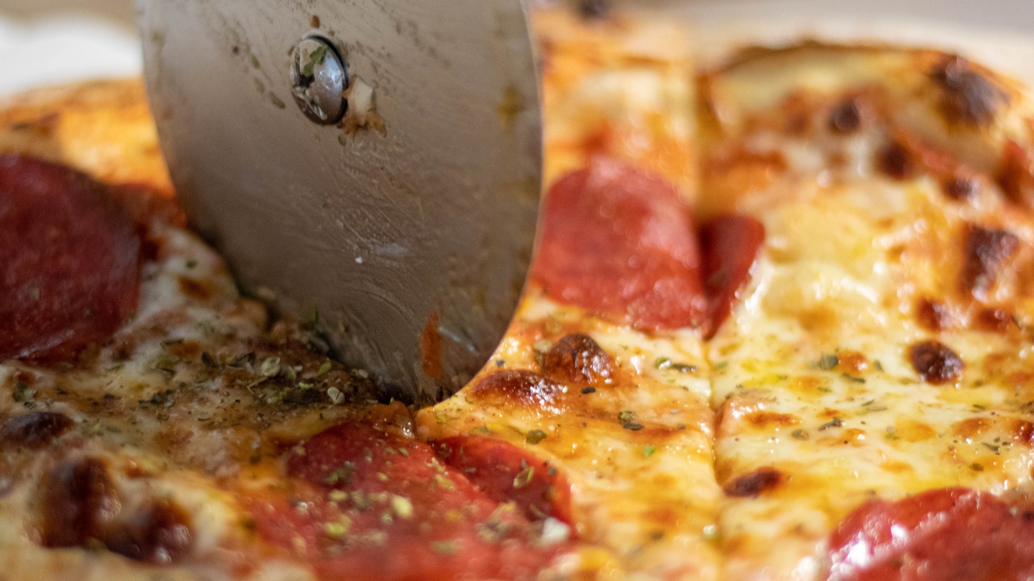 Okrągły nóż do pizzy przecina pizzę pepperoni z oregano, widoczne stopione ser i sos pomidorowy.