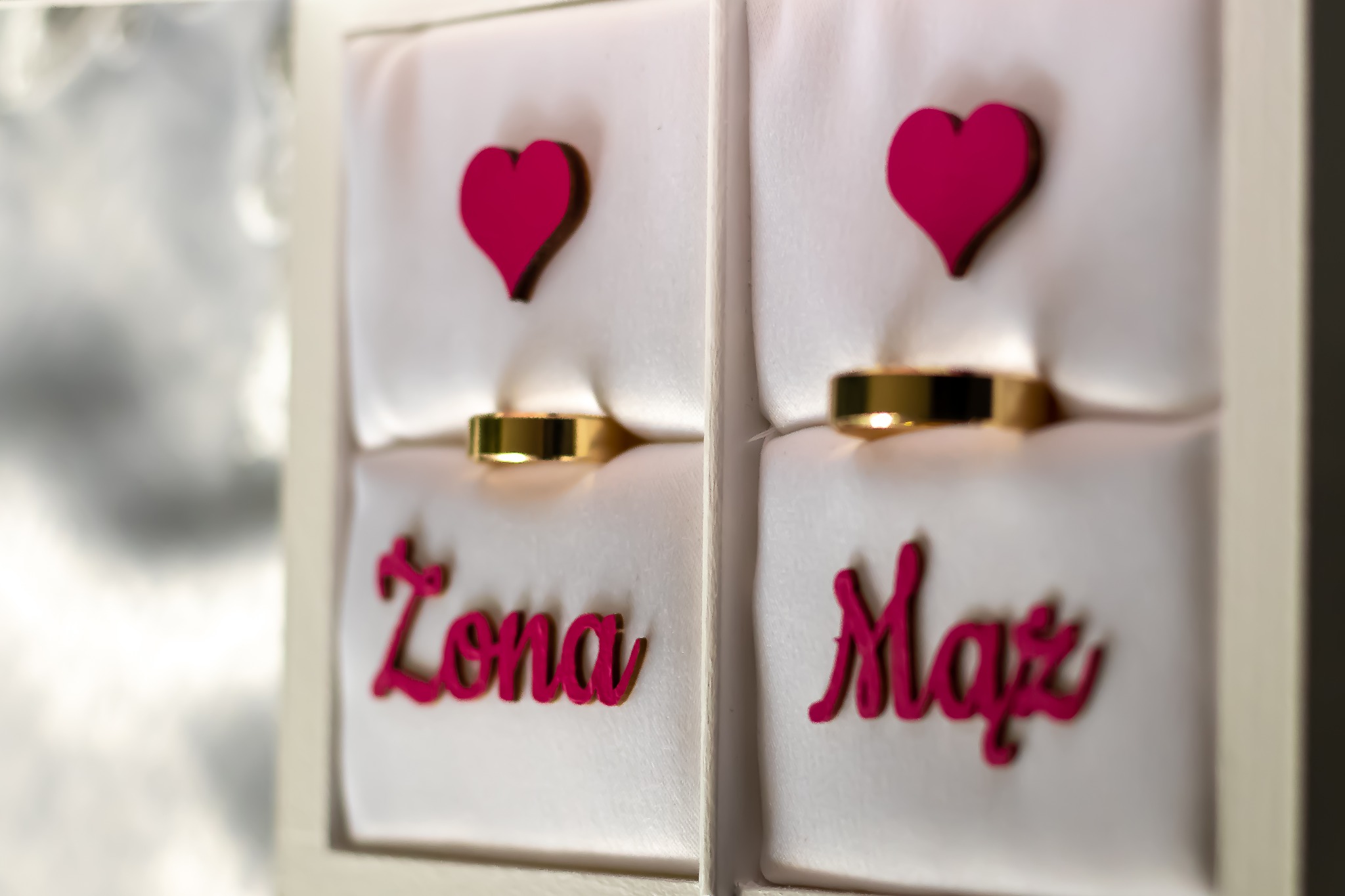 Dwa złote obrączki ślubne w białych pudełkach z napisem 'Żona' i 'Mąż' w kolorze fuksji oraz drewnianymi sercami nad nimi.