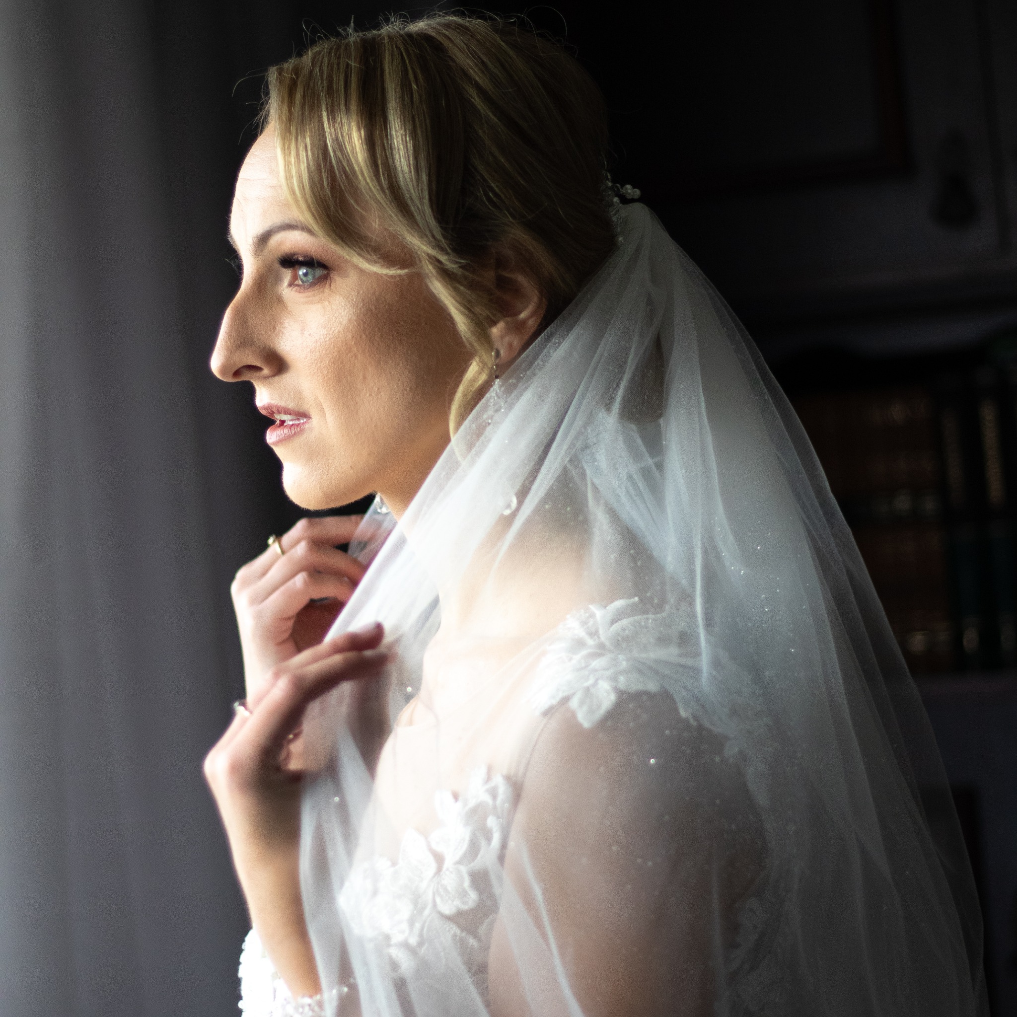 Panna młoda w białej sukni ślubnej i welonie spogląda przez okno, delikatnie dotykając welonu dłonią z pierścionkiem zaręczynowym, w tle widoczne książki.