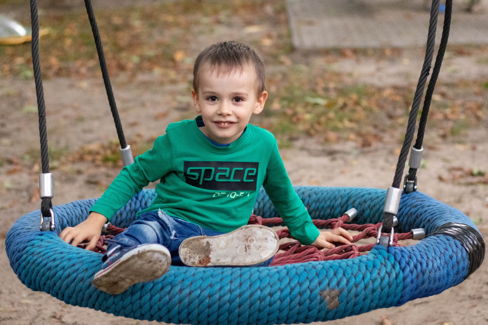 Uśmiechnięty chłopiec w zielonej bluzce z napisem 'SPACE' siedzi w okrągłej huśtawce z niebieskiej liny na tle rozmytej zieleni i brązu.