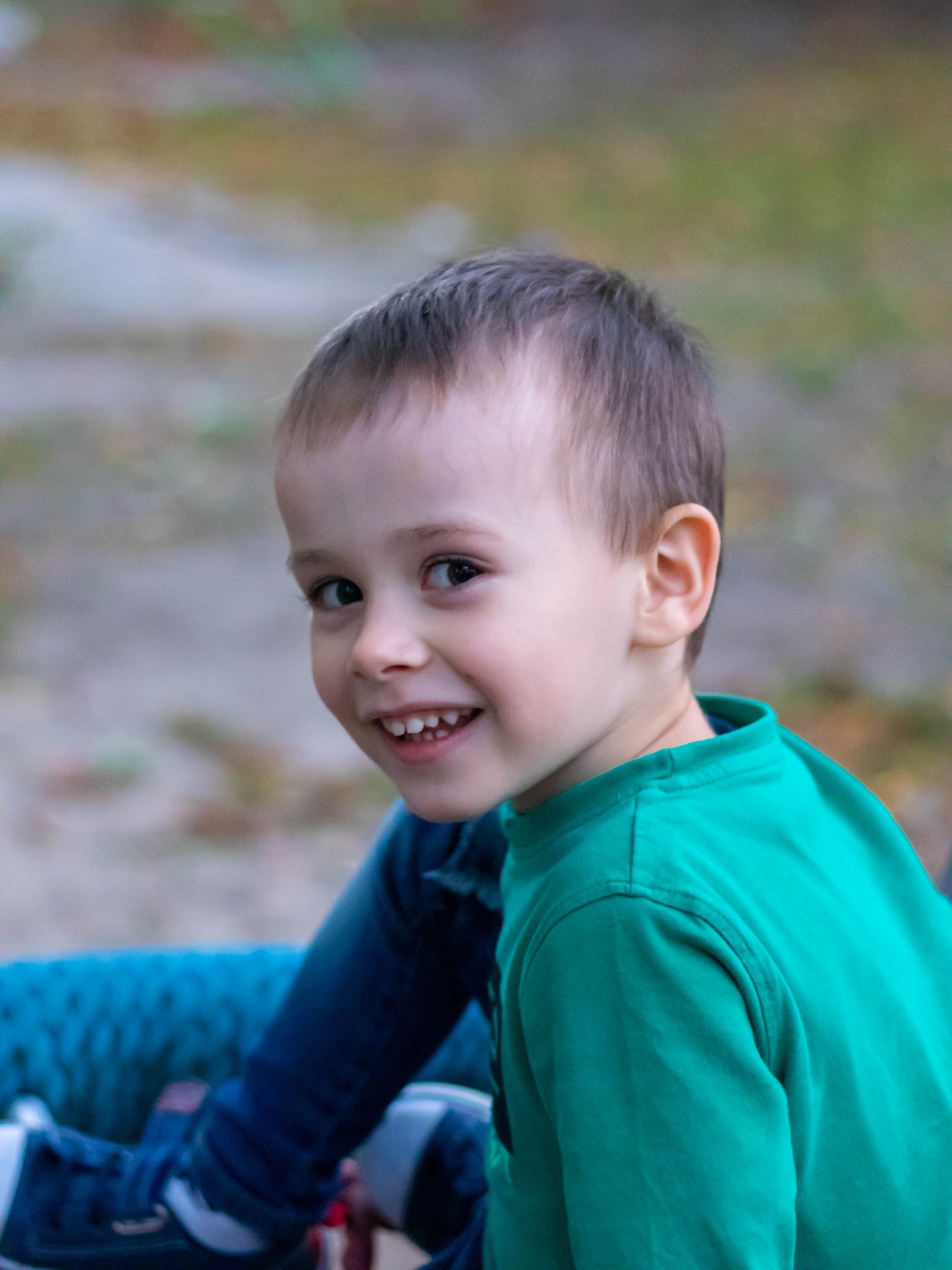 Uśmiechnięty chłopiec w zielonej koszulce siedzi, patrząc w obiektyw, z widocznymi fragmentami niebieskiego ubrania i butów w tle.