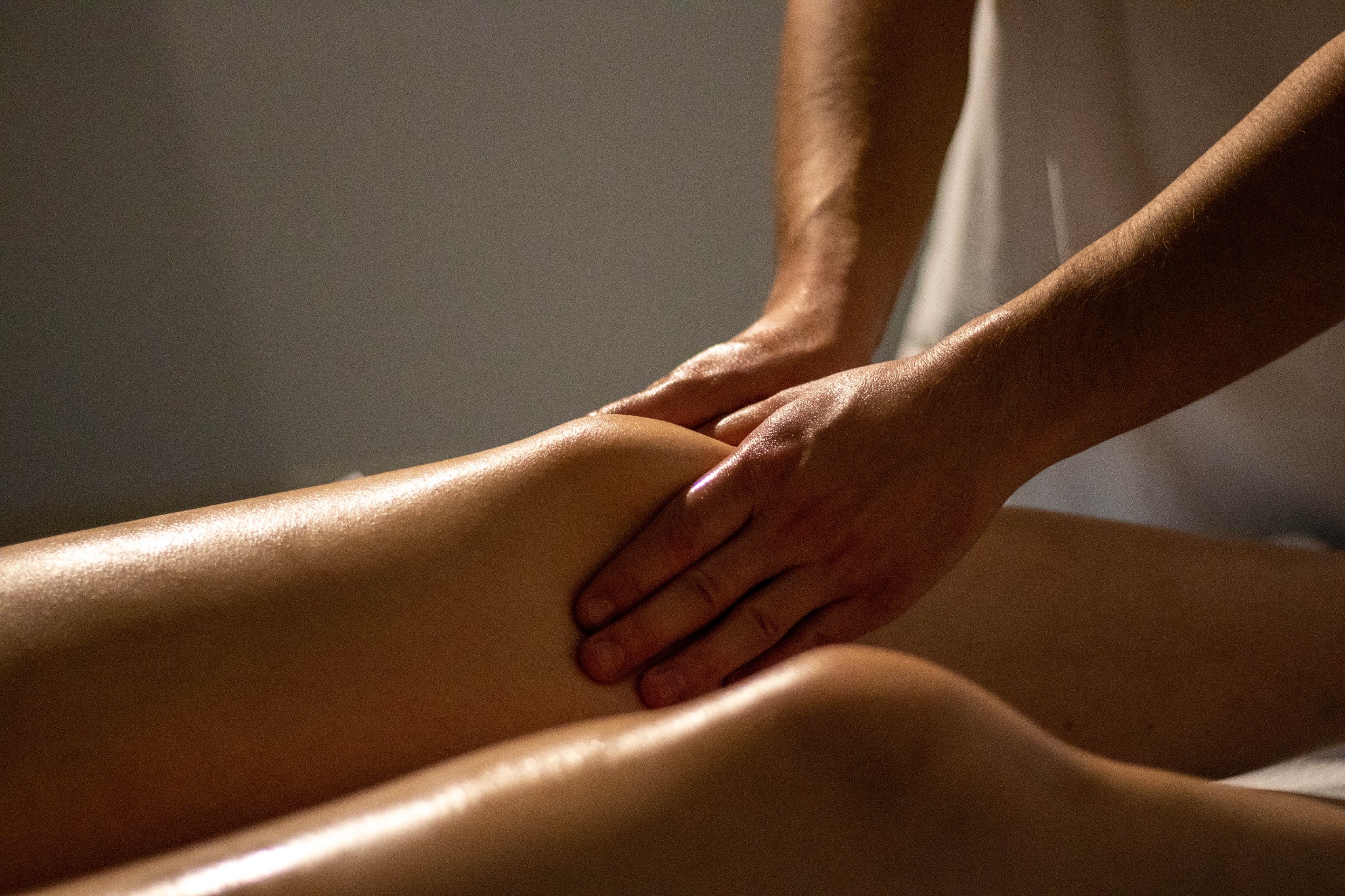 Masażysta dłońmi ugniata udo kobiety, skóra jest napięta i błyszcząca w ciepłym oświetleniu.