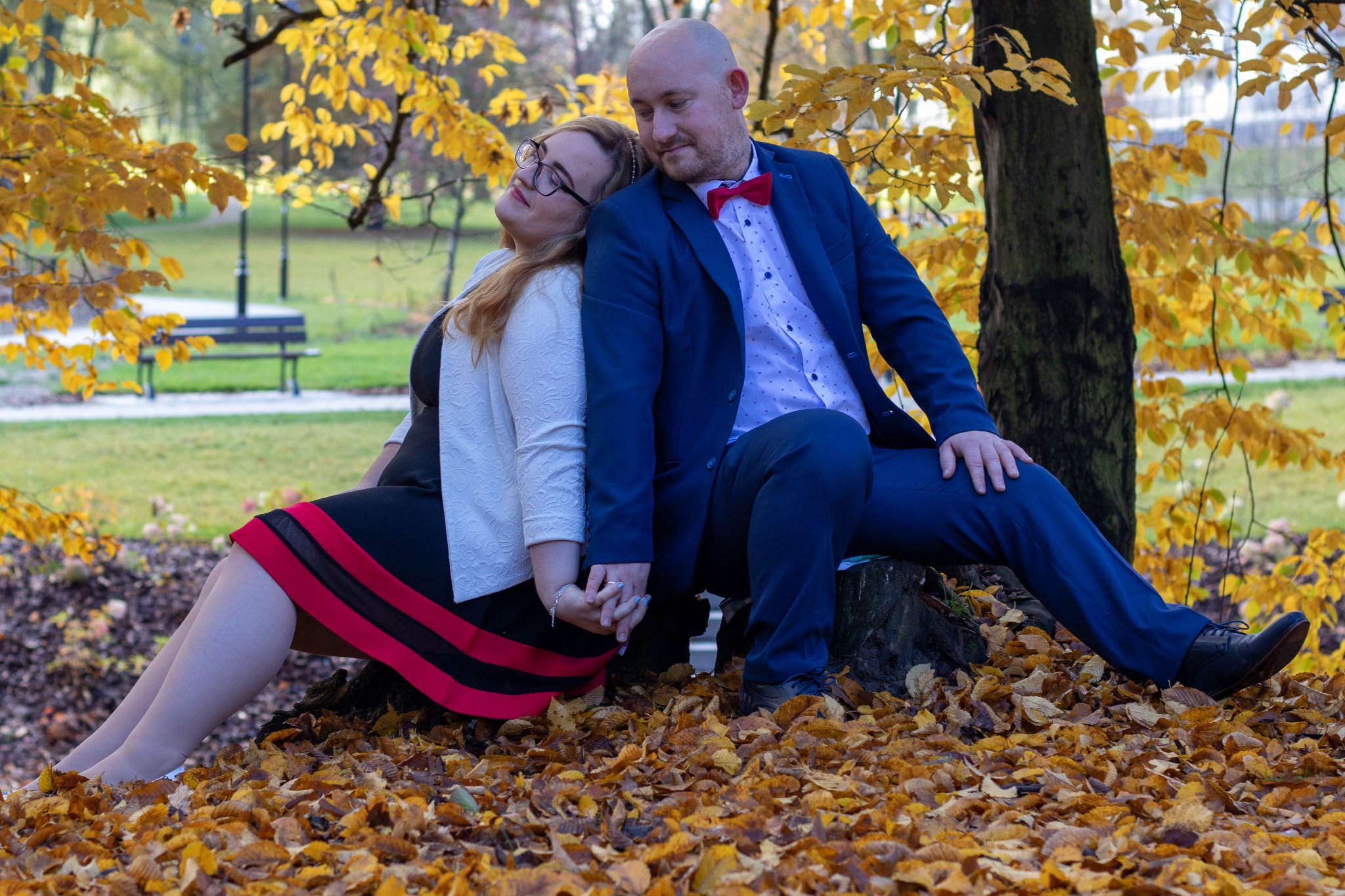 Para siedzi oparta o pień drzewa w parku jesienią, trzymając się za ręce; kobieta w czarnej sukience z czerwonymi pasami i białym żakiecie, mężczyzna w niebieskim garniturze i czerwonej muszce.
