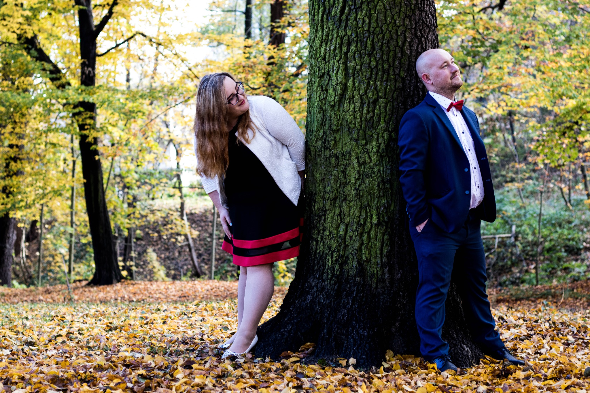 Para w jesiennym lesie pozuje przy grubym drzewie: ona w czarno-czerwonej sukience i białym żakiecie, on w granatowym garniturze i muszce, oboje uśmiechnięci.