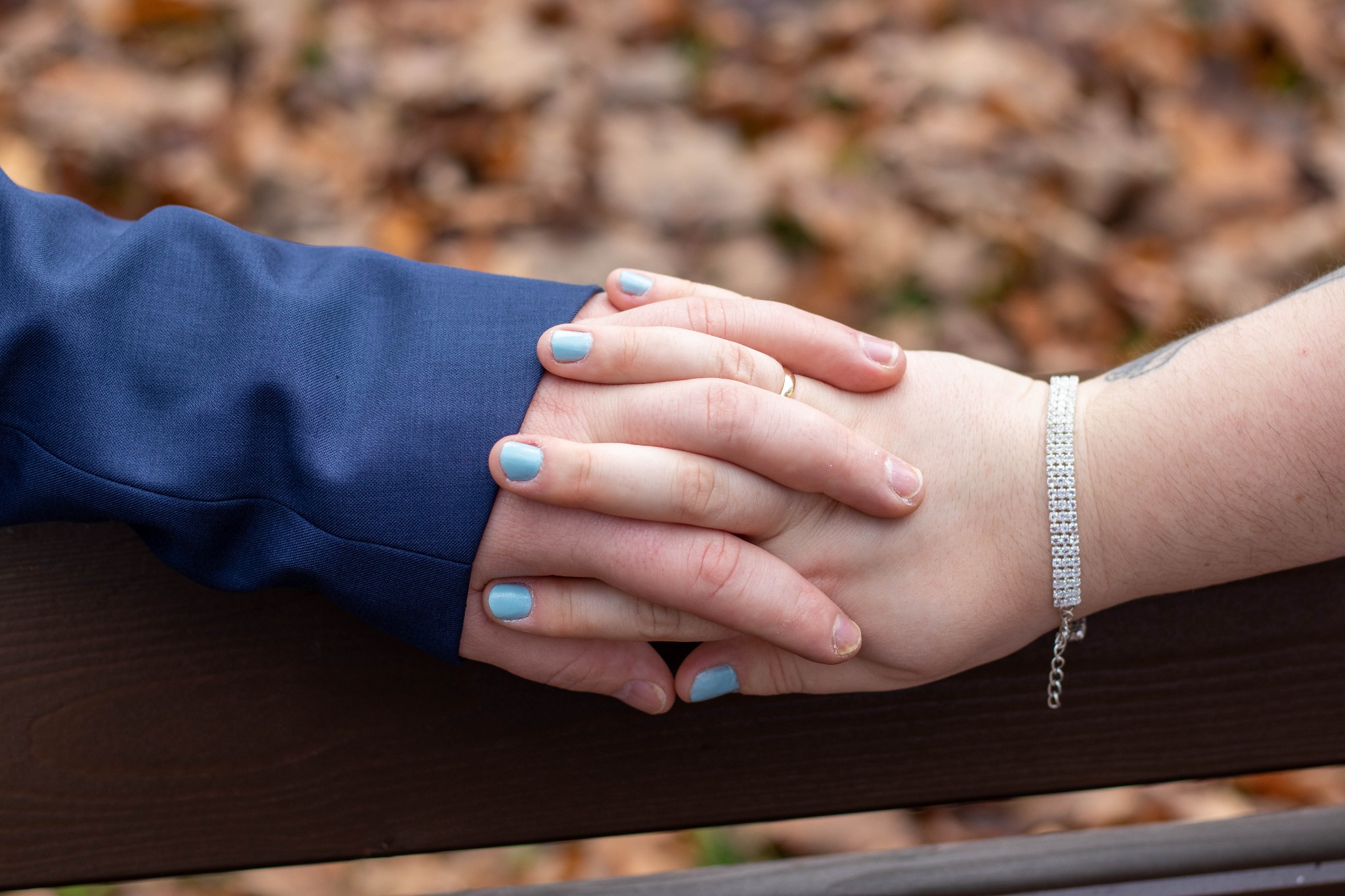 Splecione dłonie pary na tle jesiennych liści, ona z srebrną bransoletką, on w granatowym garniturze, oboje z jasnoniebieskim manicure.