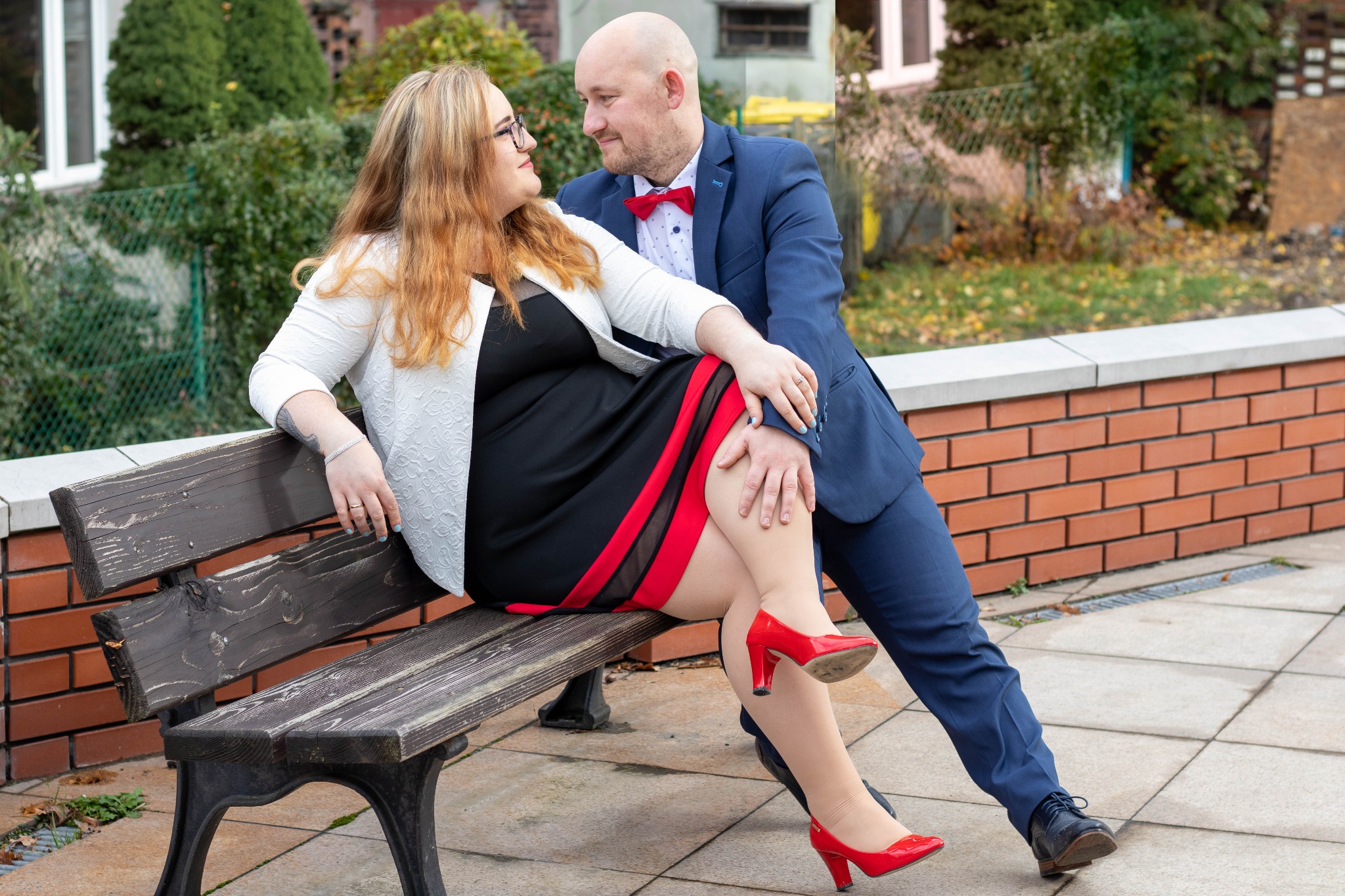 Para zakochanych: kobieta w czarnej sukience z czerwonymi akcentami i czerwonych szpilkach siedzi na ławce, opierając się o mężczyznę w niebieskim garniturze i czerwonej muszce, wymieniając...