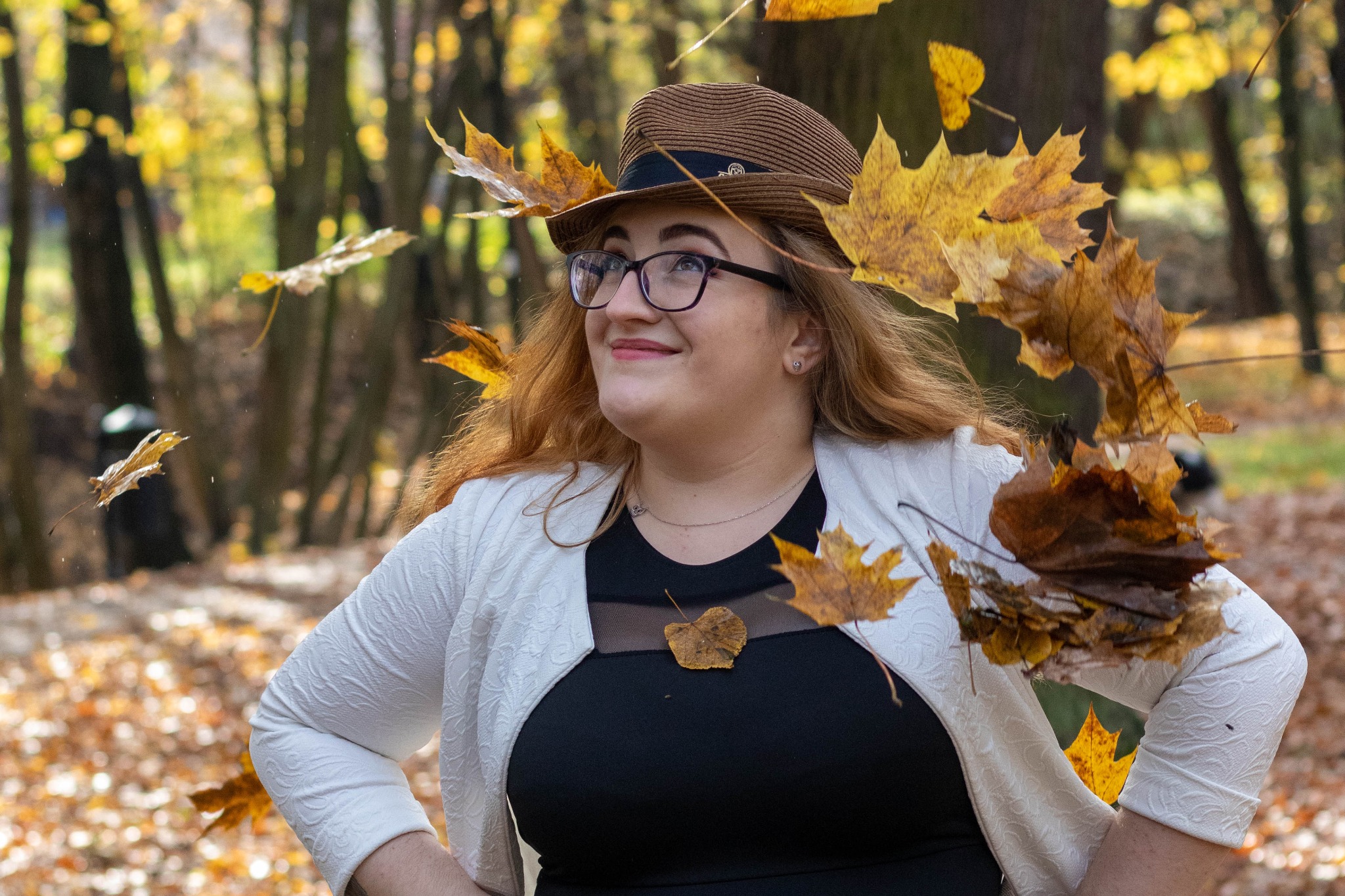 Uśmiechnięta kobieta w okularach i kapeluszu pozuje w jesiennym lesie, otoczona opadającymi liśćmi.