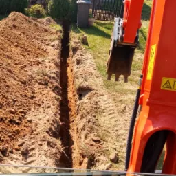 Wąski wykop w ziemi wykonany pomarańczową koparką, widok z kabiny operatora, w tle dom z szarego pustaka i rząd tuj.