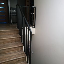 Dw-Spaw - Drewniane schody z czarną, minimalistyczną balustradą z pionowych prętów. Widok z góry na dół, z fragmentem drzwi i szafy w tle. Nowoczesny design.