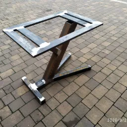 Surowa, spawana konstrukcja z metalowych profili, przypominająca ramę stołu, z widocznymi śladami spawania i szlifowania, ustawiona na brukowanej powierzchni.
